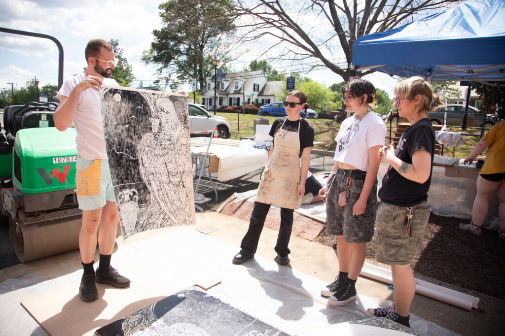 A professor holds up a print to show three students with a rented steamroller and a small tent in the background.