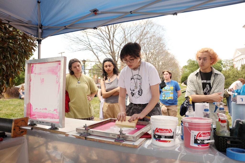 Students watch as one student screen-prints a shirt under a tent. 