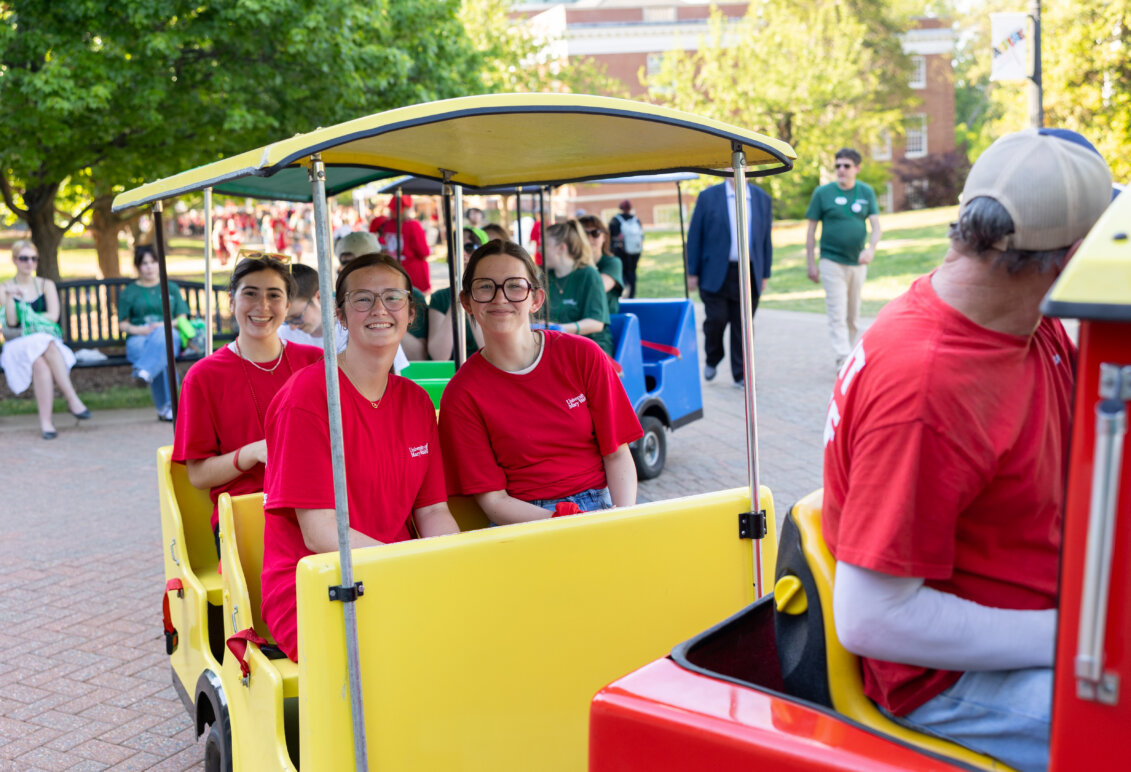 Students dressed in red ride a multi-colored train on a college campus