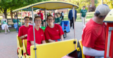 Students dressed in red ride a multi-colored train on a college campus