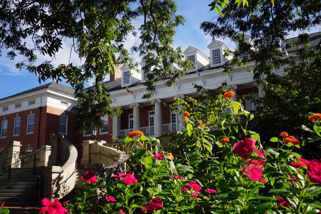 Photo of a brick building with white columns and bright pink flowers in front of it