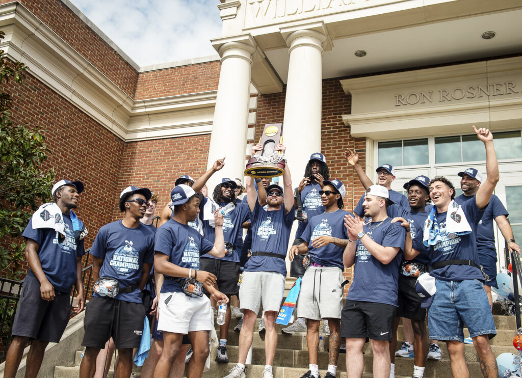A group of basketball players stand outside an arena holding a national championship trophy
