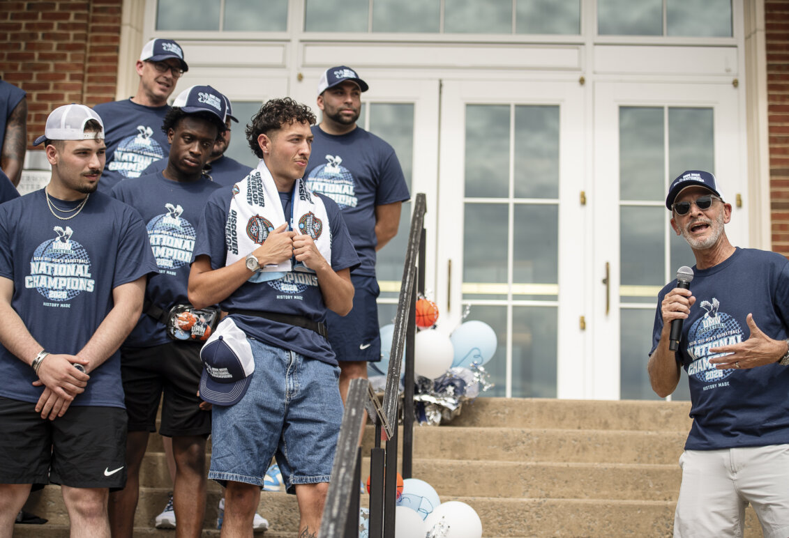 A university president speaks into a microphone to a team of basketball players on the steps of a building