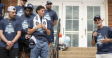 A university president speaks into a microphone to a team of basketball players on the steps of a building