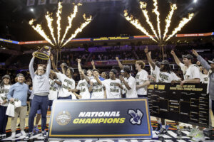 Photo of men's basketball coach and the team wearing white t-shirts that say "national champions" holding the trophy