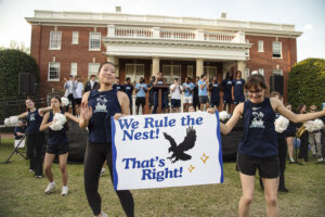 Photo of students on the lawn of Ball Circle in front of a stage with the players;a In the foreground two students from Pep Dance team hold a sign that reads "We rule the nest, that's right"