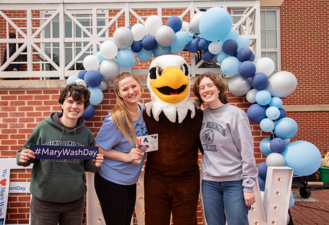 Three students stand under a blue, white and gray balloon arch with UMW mascot Sammy D. Eagle on the Cedric Rucker University Center patio.