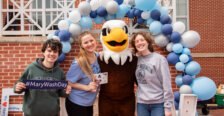 Three students stand under a blue, white and gray balloon arch with UMW mascot Sammy D. Eagle on the Cedric Rucker University Center patio.
