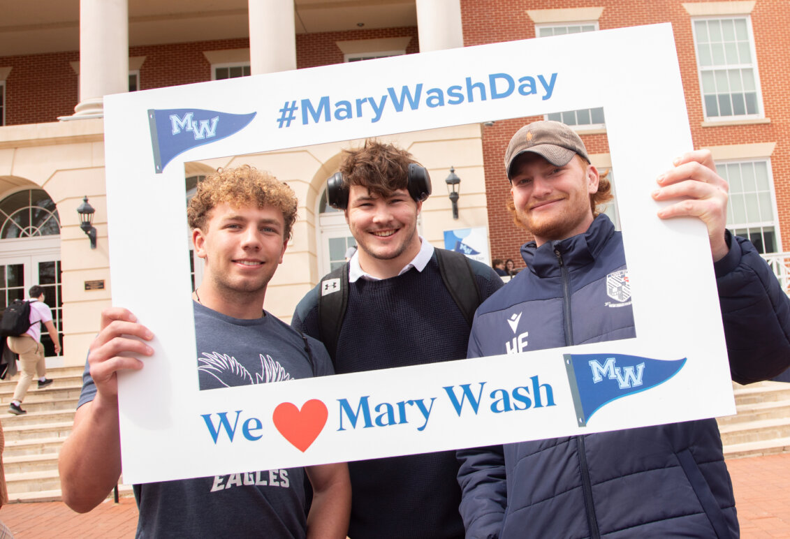 Three students in front of the Cedric Rucker University Center holding up a frame that reads #MaryWashDay and We love Mary Wash.