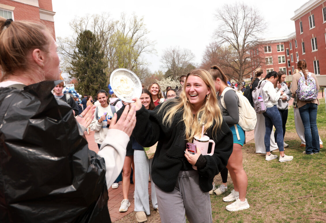 A student pies a volunteer in the face while onlookers have fun in the background.