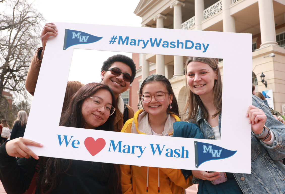 A group of four students in front of the Cedric Rucker University Center holding up a frame that reads #MaryWashDay and We love Mary Wash.