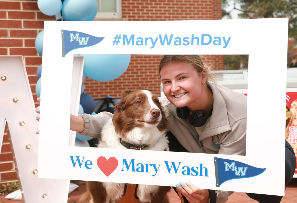 A student and her dog pose for a picture with a sign that says #MaryWashDay and We love Mary Wash.