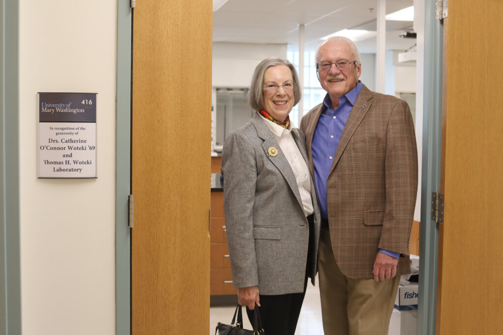 A couple standing in the doorway of a laboratory with their names on the plaque.