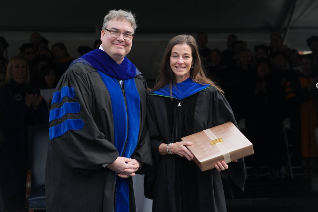 A man and a woman dressed in graduation garb smile for the camera, while the woman holds a box
