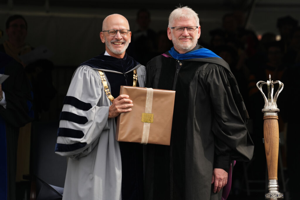 Two men dressed in graduation garb smile for the camera, while one holds a box