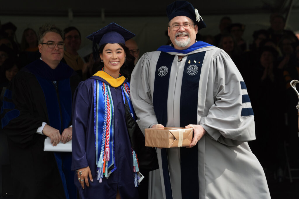 A young woman and a man dressed in graduation garb stand together, while the man holds a box