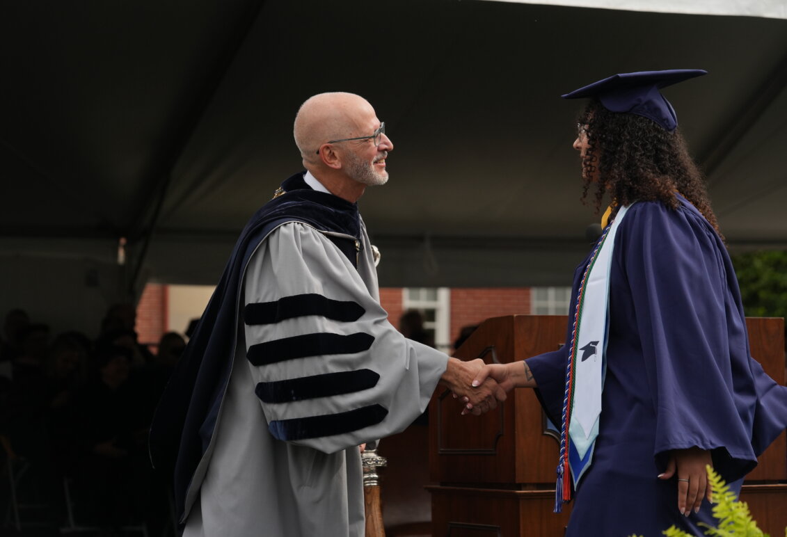 A man and a woman dressed in graduate garb shake hands onstage
