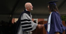 A man and a woman dressed in graduate garb shake hands onstage