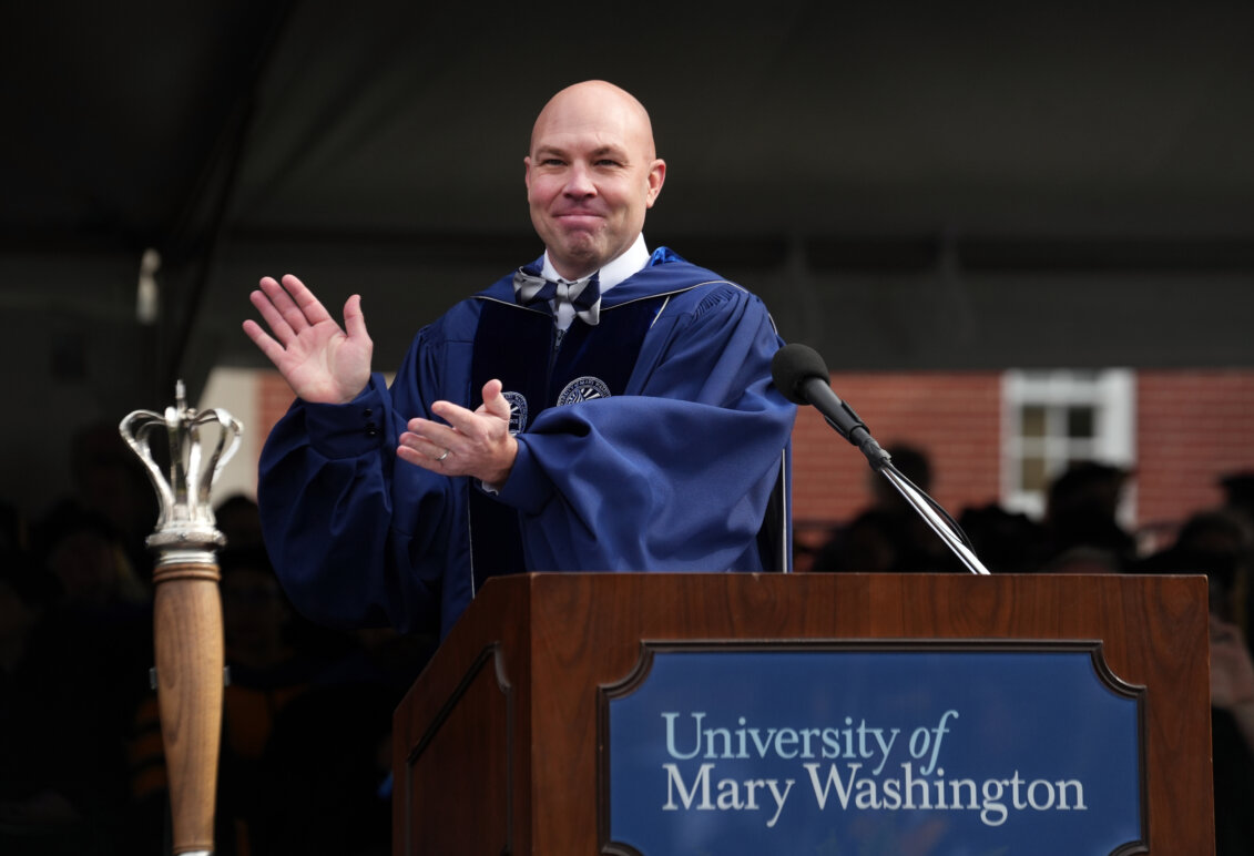 A man dressed in graduation garb stands behind a podium with the ceremonial mace in the foreground