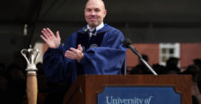 A man dressed in graduation garb stands behind a podium with the ceremonial mace in the foreground