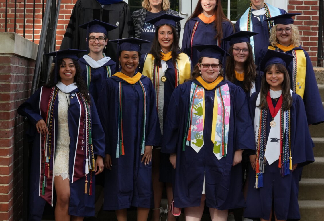 A group of students and faculty, mostly dressed in graduation garbs, stand on the steps of a brick building