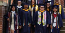 A group of students and faculty, mostly dressed in graduation garbs, stand on the steps of a brick building