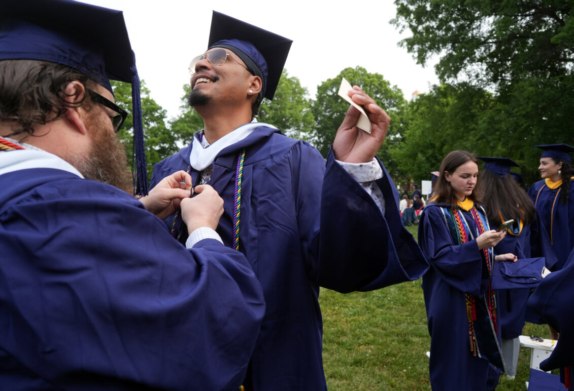 Two guys dressed in graduation garb, with one of them helping adjust the other's cords