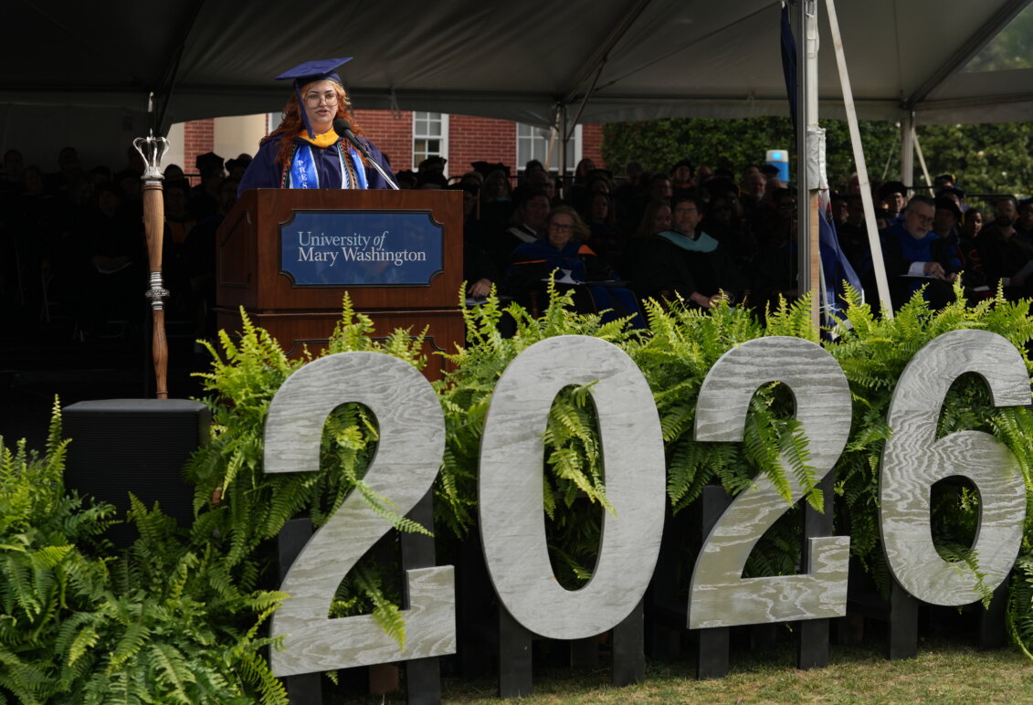 A girl dressed in graduation garb stands behind a podium with the number "2026" in the foreground