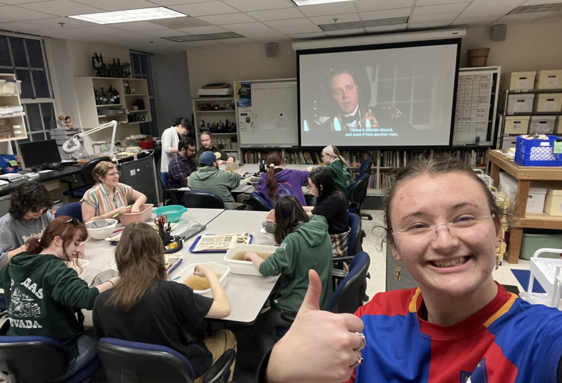 Bowman gives the camera a thumbs up with students in the background are at tables in a classroom and movie is playing on a projection screen