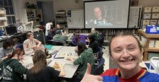 Bowman gives the camera a thumbs up with students in the background are at tables in a classroom and movie is playing on a projection screen