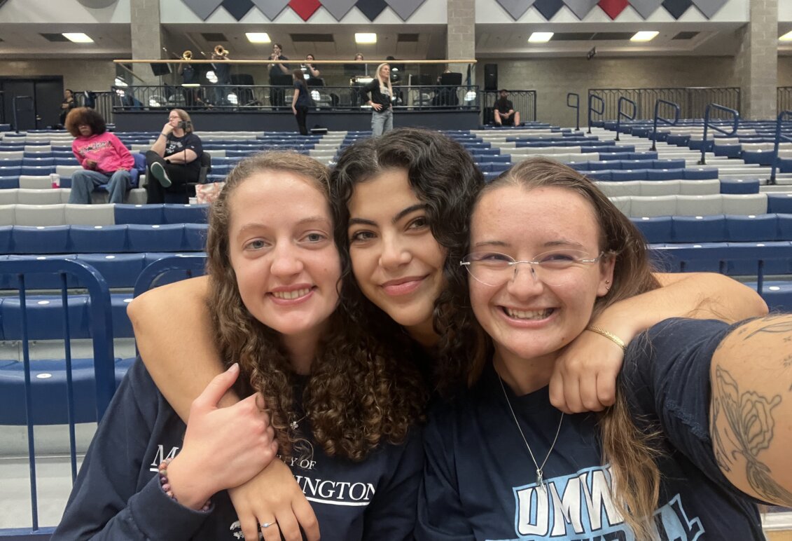 Three women smiling together in the Ron Rosner arena on campus
