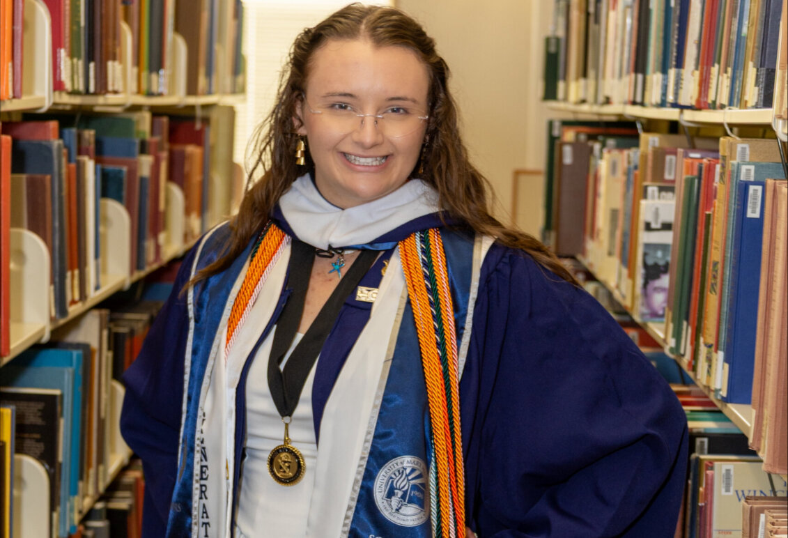 Makayla Bowman standing in the library aisle with graduation robes and cords