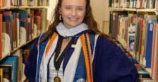 Makayla Bowman standing in the library aisle with graduation robes and cords