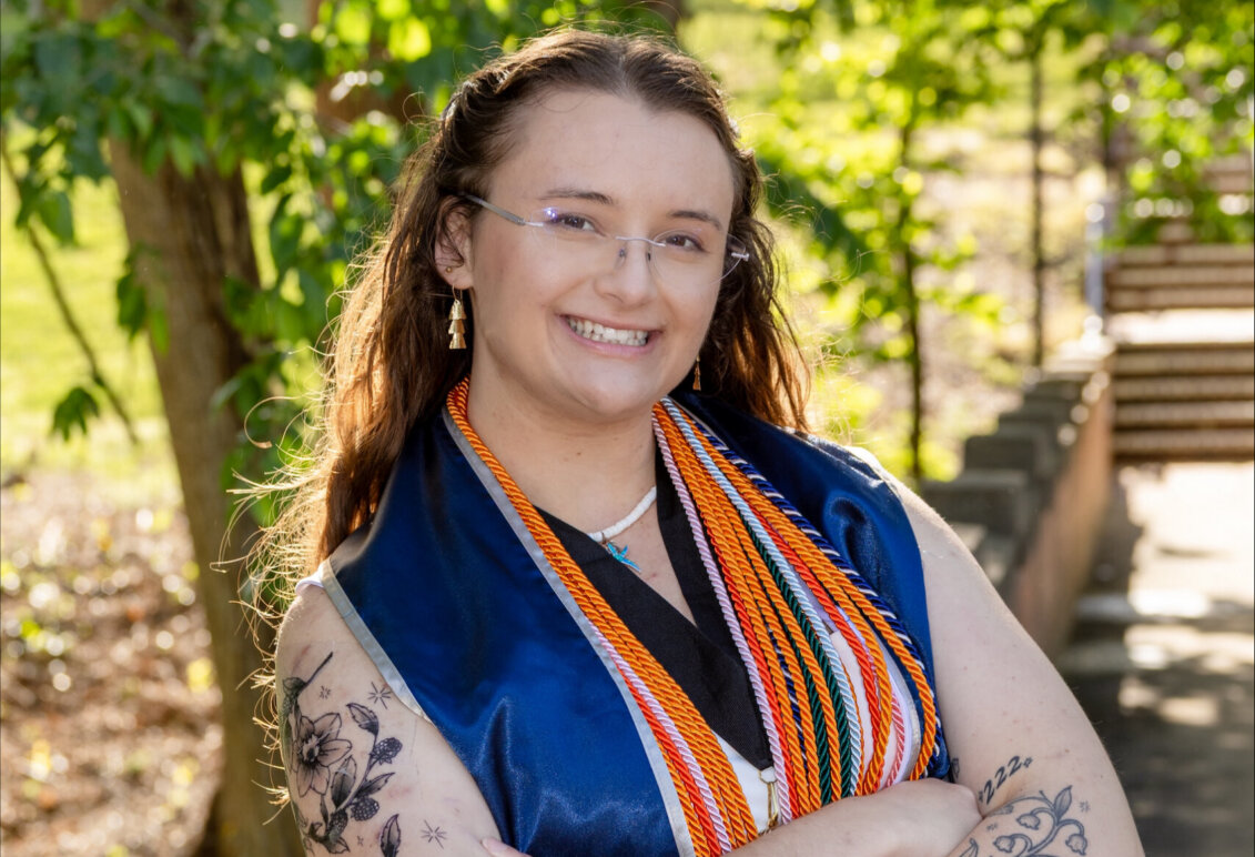 Bowman poses outdoors on campus with graduation robe and cords