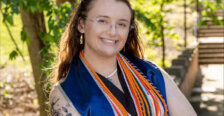 Bowman poses outdoors on campus with graduation robe and cords