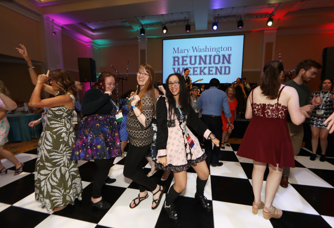 Alumni dance on a black-and-white checkered floor with a screen that reads 'Mary Washington Reunion Weekend' behind them. The room has bright colored lights around to make it festive.