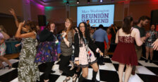 Alumni dance on a black-and-white checkered floor with a screen that reads 'Mary Washington Reunion Weekend' behind them. The room has bright colored lights around to make it festive. 