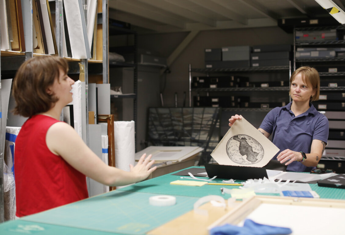 A professor holds a piece of artwork up while discussing it with a student.