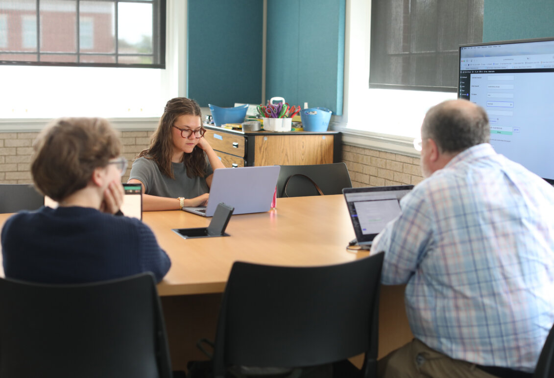 A professor and two students sit at a classroom table with laptops.