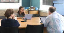 A professor and two students sit at a classroom table with laptops.