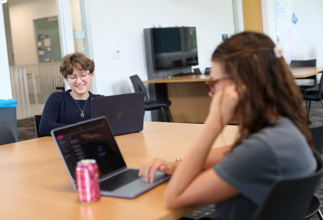 Two students sit at a laptop, while one looks at a laptop.