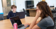 Two students sit at a laptop, while one looks at a laptop.