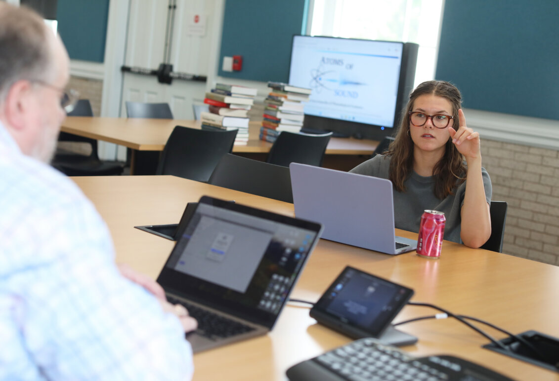 A student sits across the table from a professor, while they are both on their laptops.