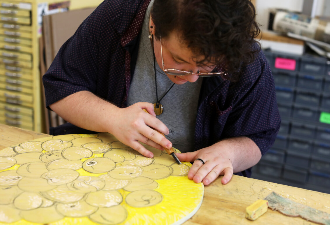 An art student carves away at a round, yellow piece of wood.