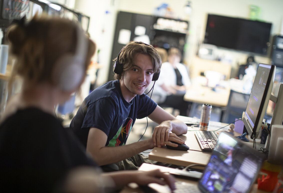Two students wearing headphones talk in front of their laptops.