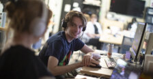 Two students wearing headphones talk in front of their laptops.