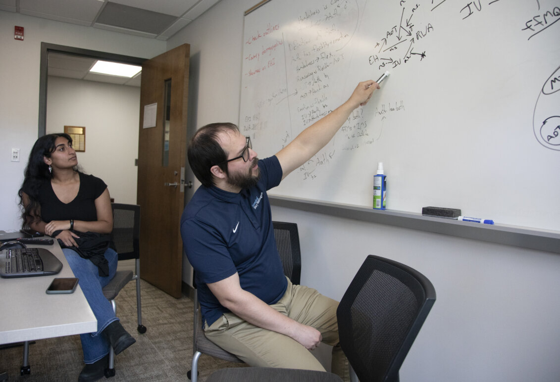 A professor points to an equation on a whiteboard while a student looks on.