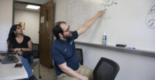 A professor points to an equation on a whiteboard while a student looks on.
