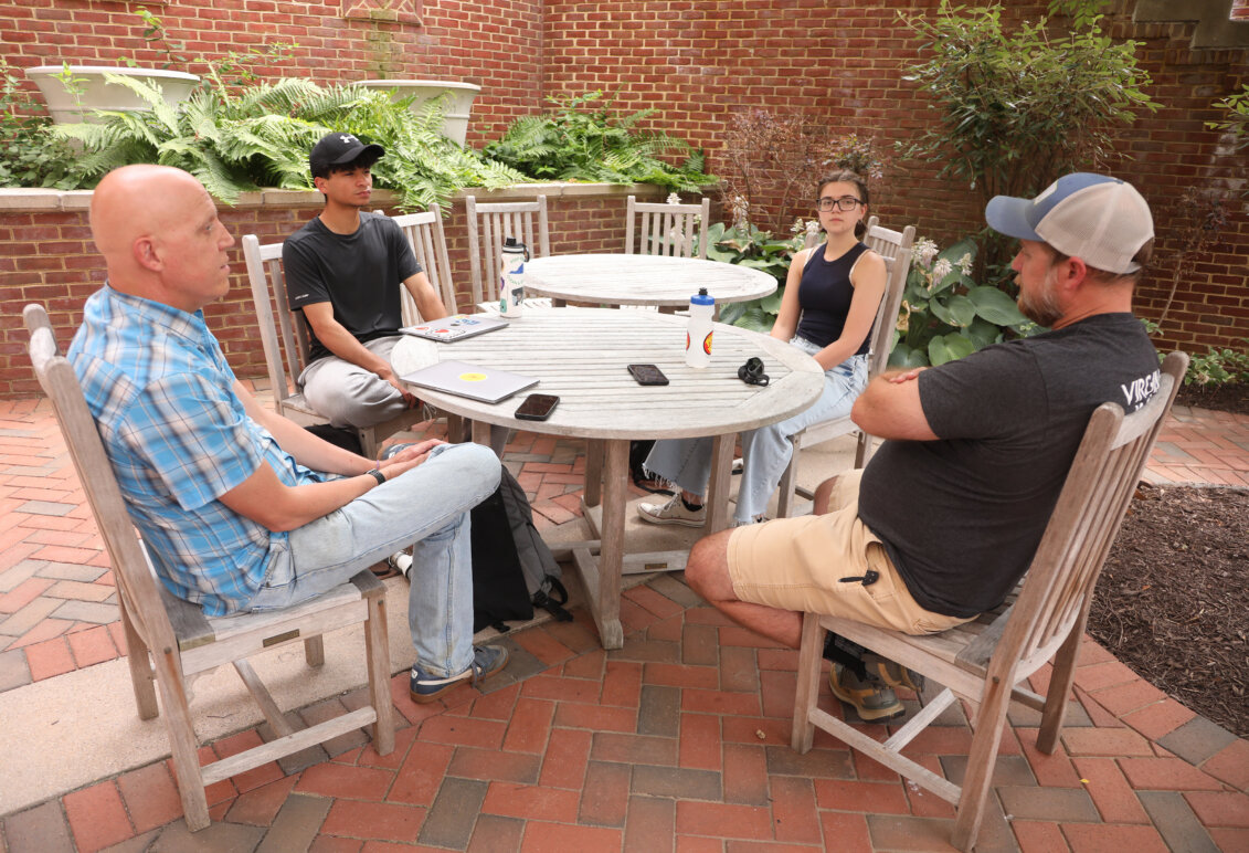 A professor, an expert on the Rappahannock River and two UMW students sit around a table near a brick building on campus.