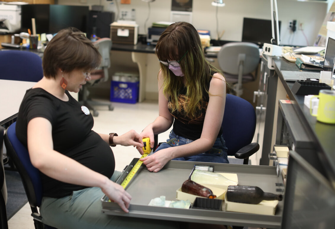 A professor and a student sit in a historic preservation lab and hold a measuring tape against a storage drawer.